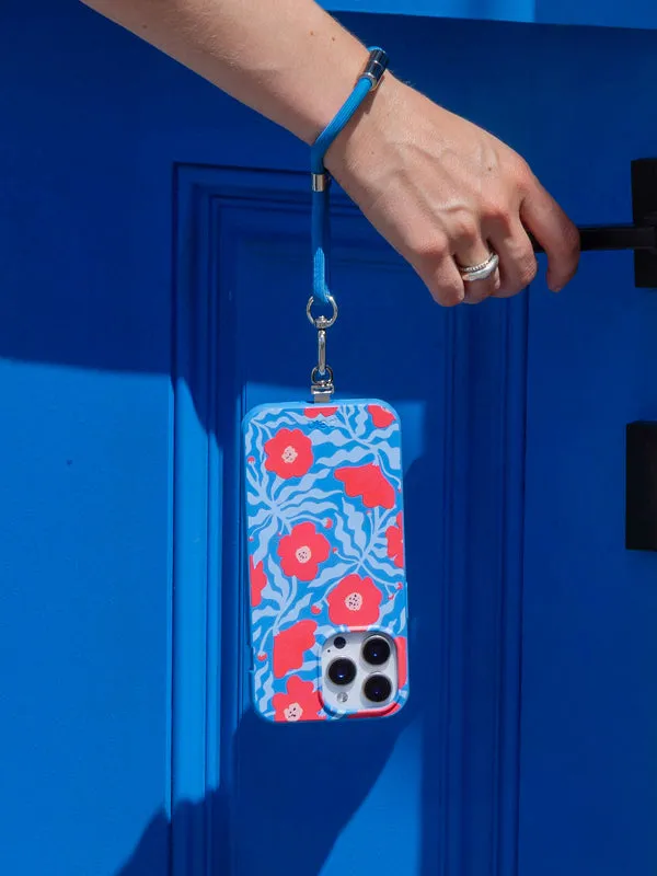 A hand holding a blue floral Pela phone case on a strap against a blue door