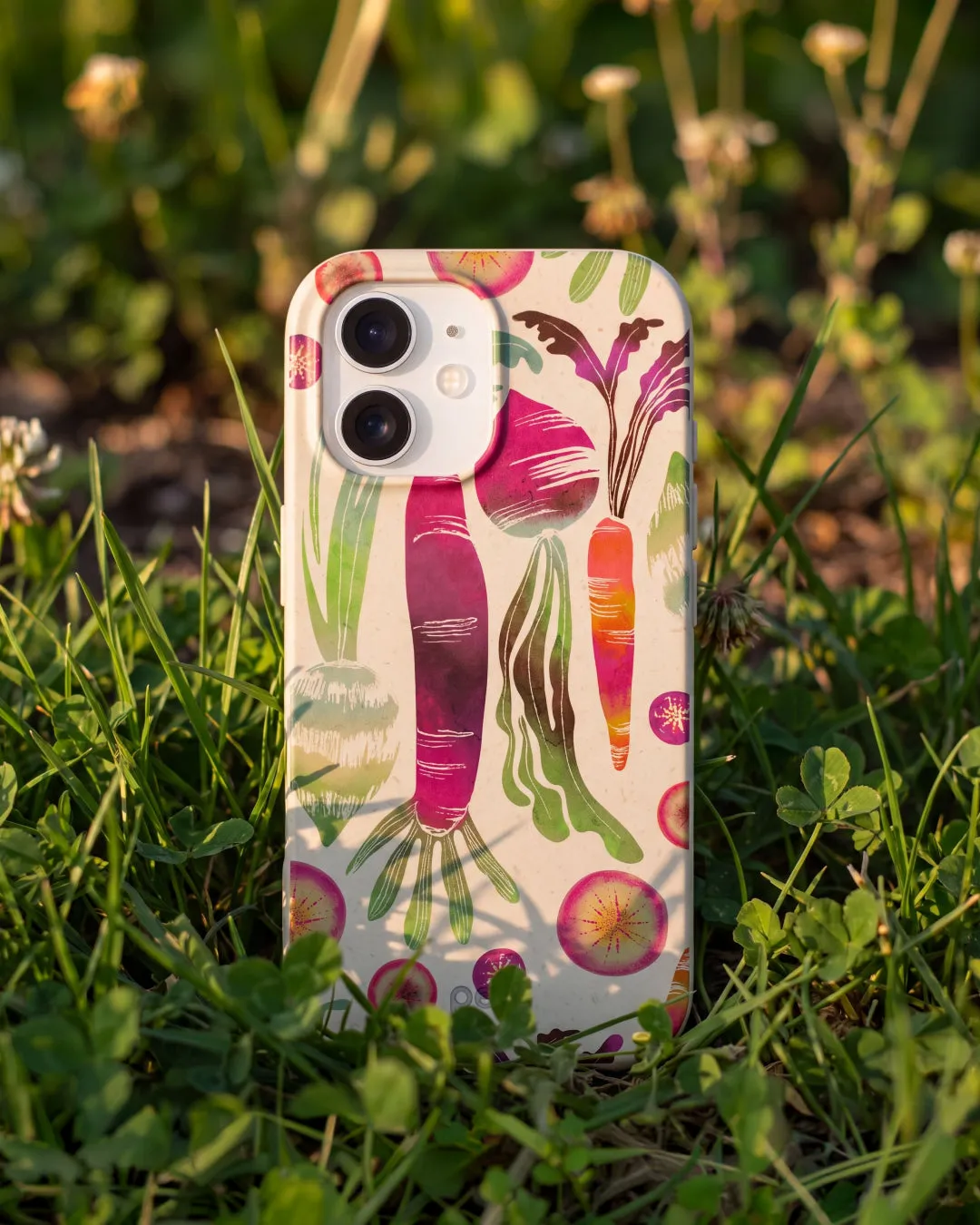 A Pela Root Groove phone case in London Fog featuring bold illustrated root vegetables — beets, carrots, radishes, and greens in rich magenta, orange, and green tones — standing upright in lush green grass among clover and wildflowers in warm golden hour sunlight.