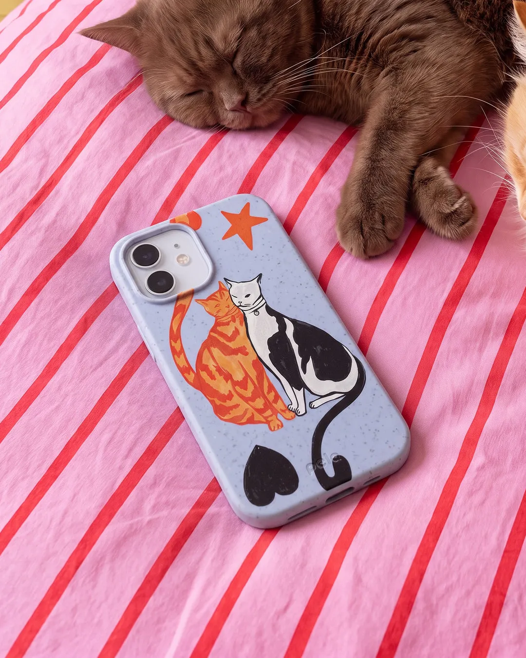 A Pela Twins phone case in Powder Blue featuring two illustrated cats — an orange tabby and a black and white cat — nuzzling together with a red star and black hearts, resting on a pink and red striped bedsheet beside a sleeping brown cat.
