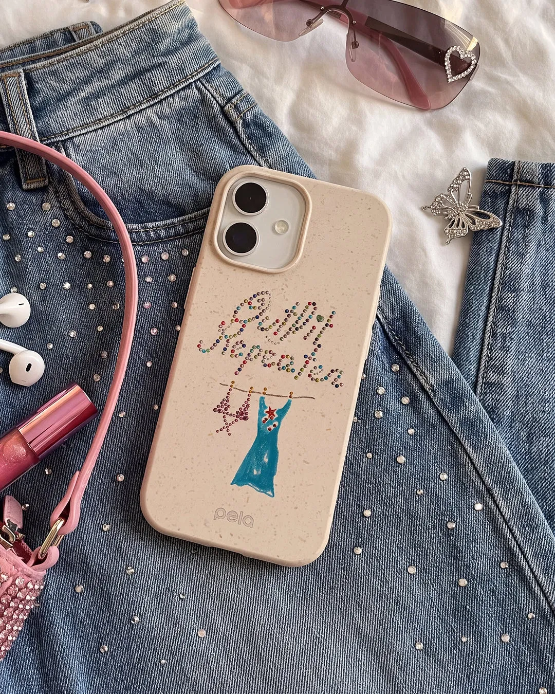 A Pela Outfit Repeater phone case in Seashell featuring rhinestone-style lettering and an illustrated blue dress on a hanger, styled on a rhinestone-studded denim jacket alongside a pink phone strap, white earbuds, pink lip gloss, rimless tinted sunglasses, and a crystal butterfly brooch.