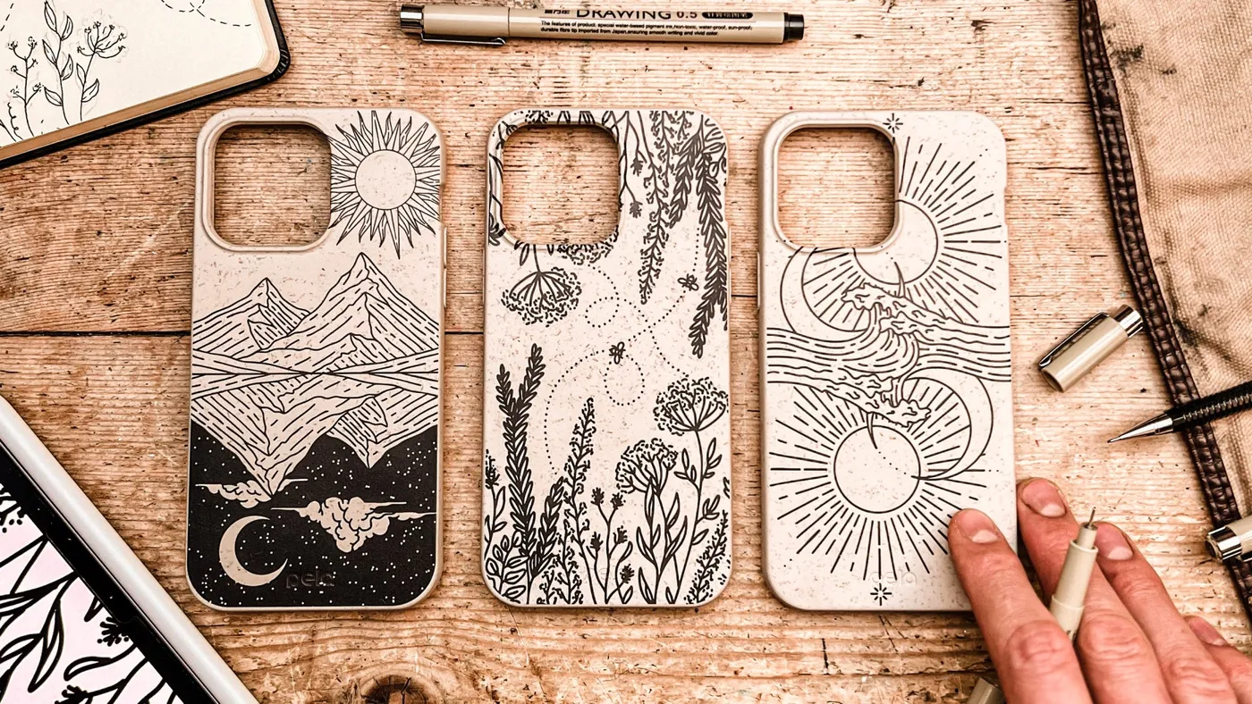 Three Pela phone cases with engraved line-art designs on a wooden desk beside sketches